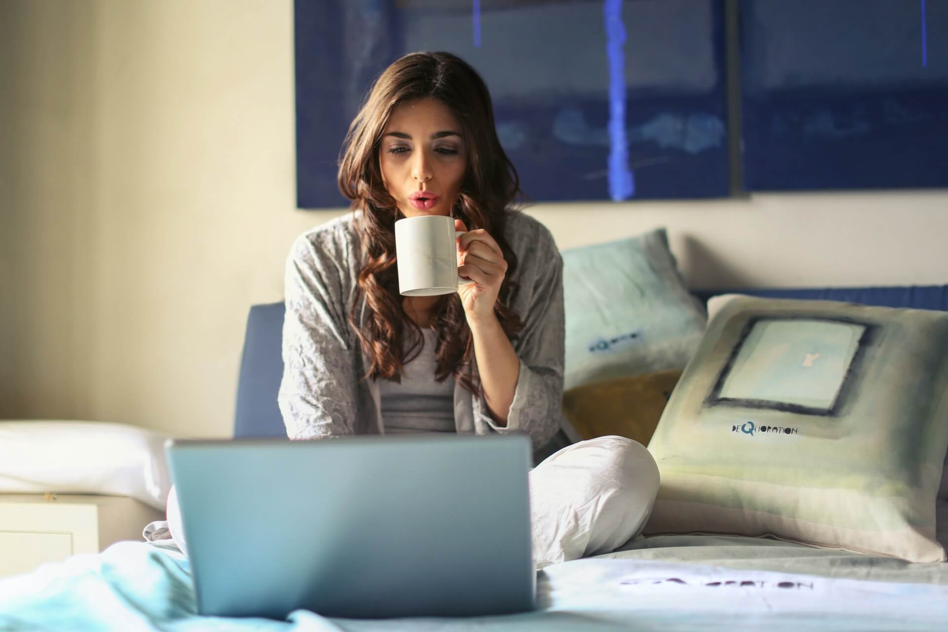 Woman sitting on bed using laptop and drinking coffee using an online chat room.